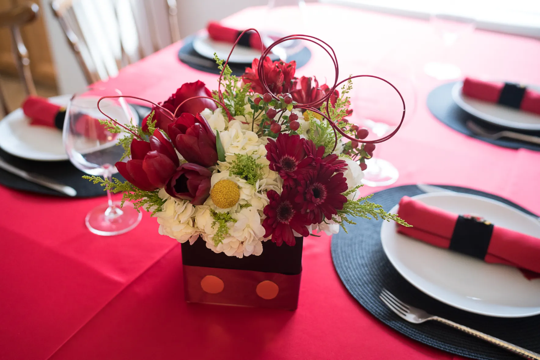 Baby-Shower Baby shower and gender reveals table centerpiece with red tulips, burgundy gerbera, white hydrangea, and curly willow accents in a polka dot box vase on a red linen table