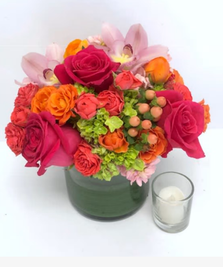 Bouquet of roses in hot pink, coral, and orange with green hydrangeas, pink orchids, and berries in a dark green glass vase, displayed next to a small white candle.