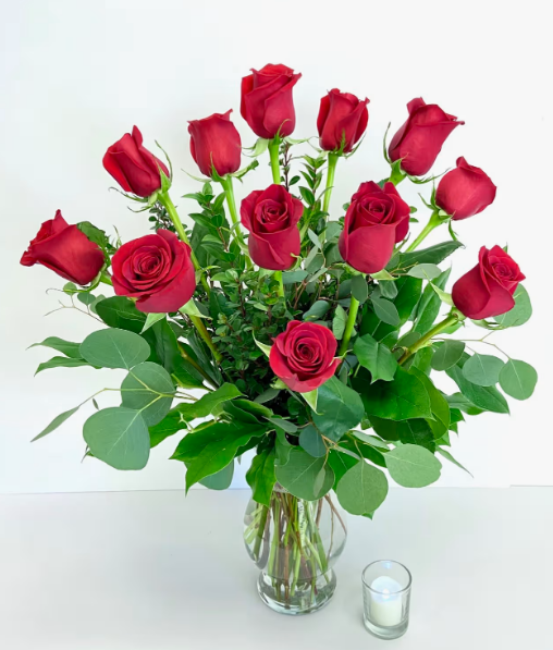 Classic dozen red rose bouquet with eucalyptus greenery arranged in a clear glass vase, displayed next to a small white candle